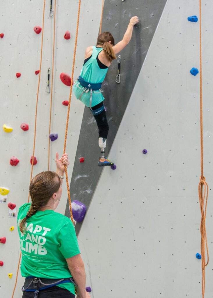 A young woman who has a prosthetic right leg climbs a wall at an indoor rock climbing gym while another woman in a green shirt belays her.