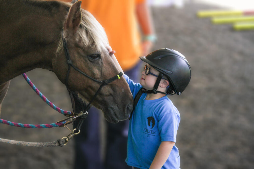 A young boy in a blue shirt wearing a helmet pets the nose of a horse who is gently extending his nose at the young boy.