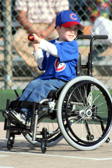 A young boy in a wheelchair wearing a Cubs jersey and hat swings a bat.