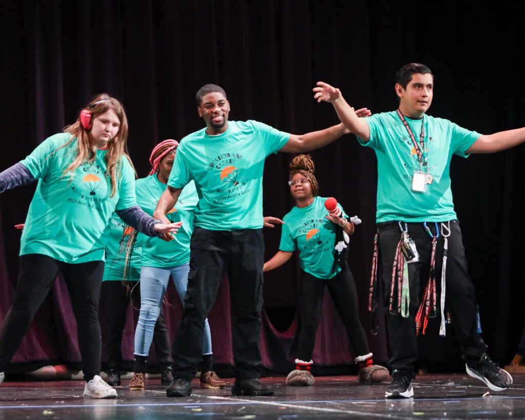 Three young women and two young men all dressed in green t-shirts and black pants dance on a stage.