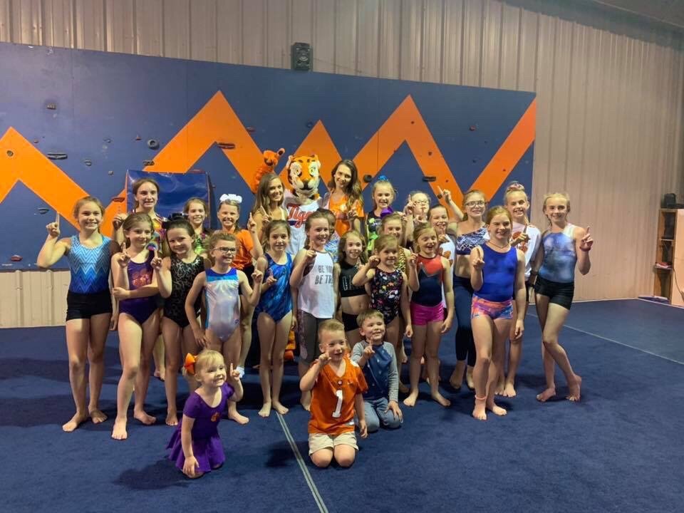 A large group of young gymnasts pose on a blue mat with the Clemson Tiger mascot.