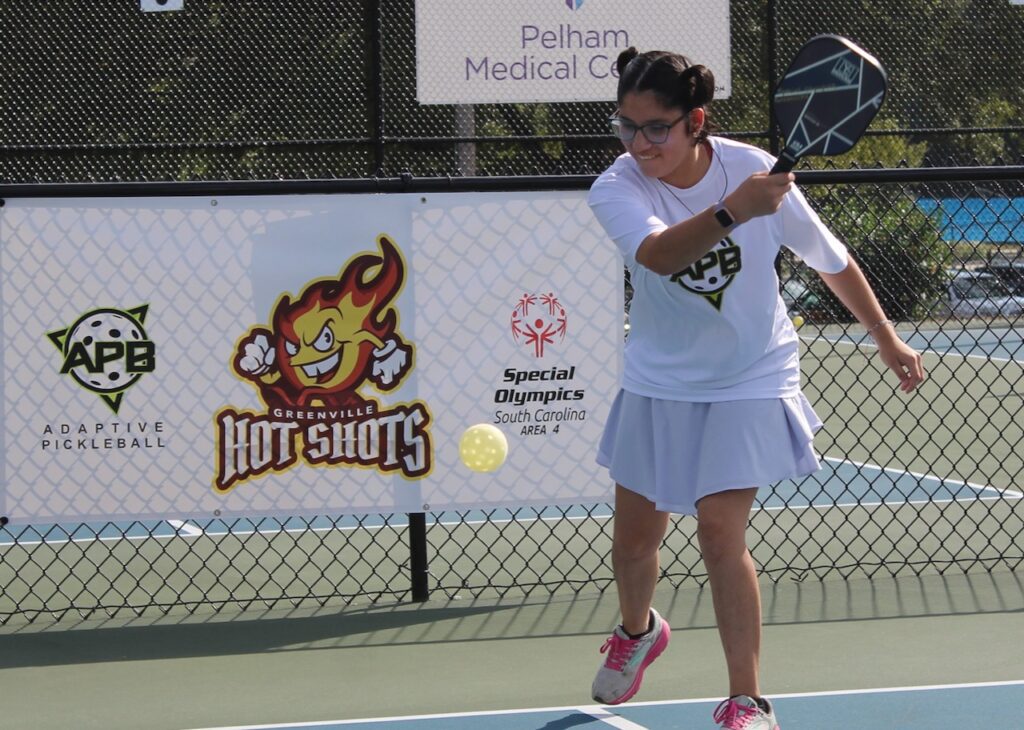 A young woman wearing a white t-shirt and a white tennis skirt swings at a pickleball on a sunny day at a Pickleball court.