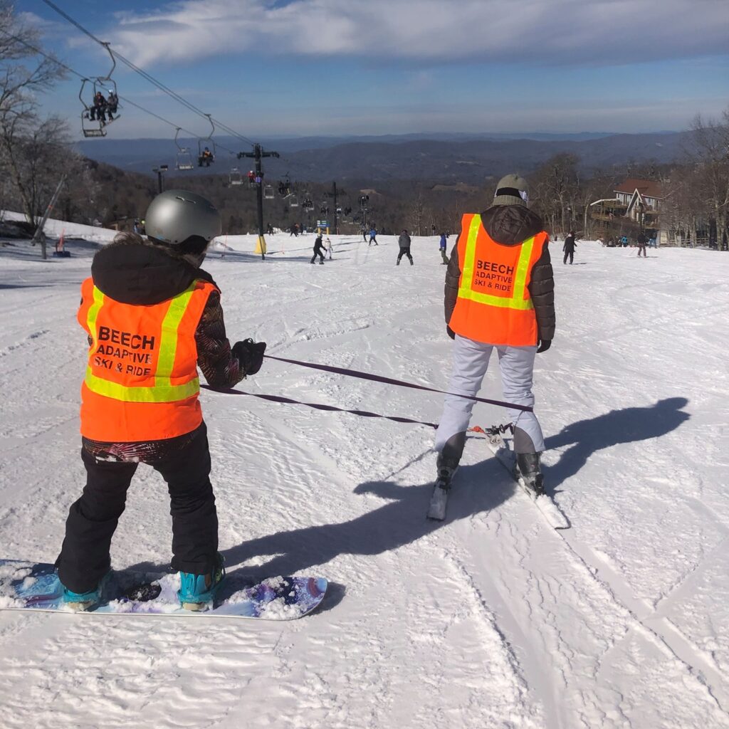 An instructor wearing a bright orange vest  uses straps to help control a visually impaired skier who is also wearing a bright orange vest.
