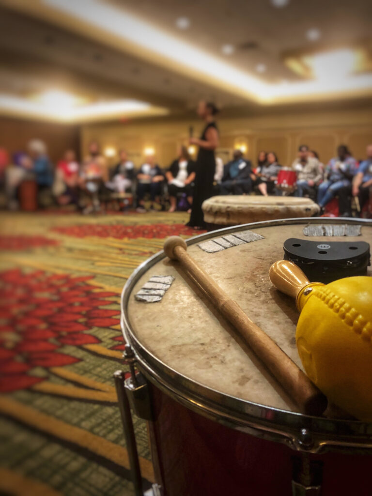 A drum is in focus in the foreground, and a large group of people sit in a circle around an instructor.
