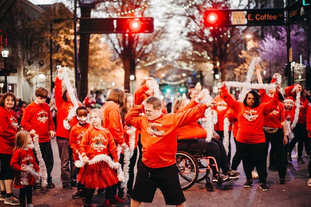 A large group of dancers wearing red and holiday themed clothes dances in the middle of Main Street Greenville, SC.
