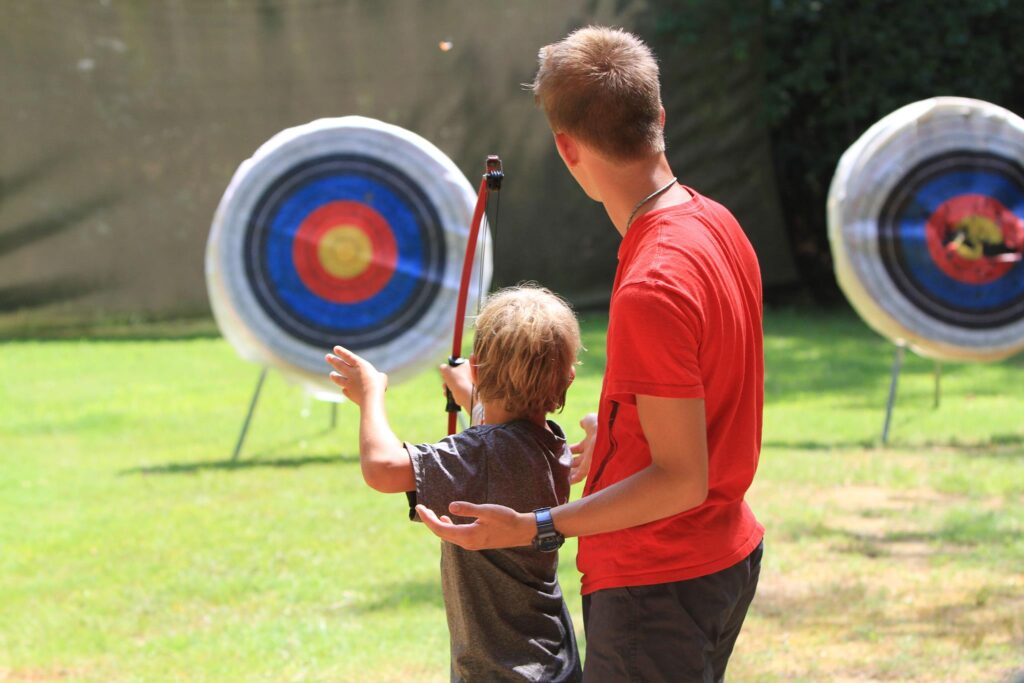 Young man shoots an arrow from a bow at a target while an adult watches.