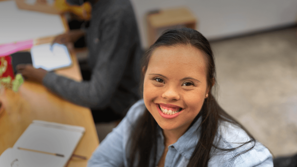 A young woman sitting at a table looks up at the camera with a smile on her face.