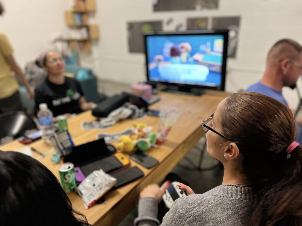 Two young women sit around a table playing a video game on a screen.