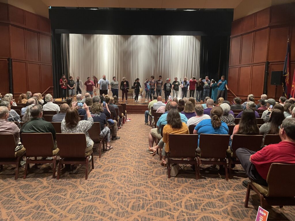 A group of people stand on stage in a line, with a audience watches them.