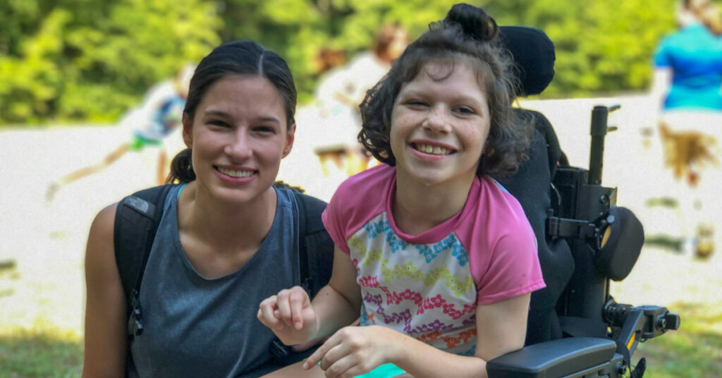 A camper sits smiling her wheelchair and holds the hand of a camp counselor.