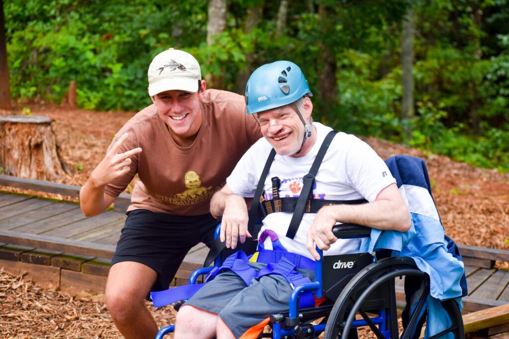 Two men smile for the camera, one of the men is wearing a helmet and preparing to climb the confidence tower.