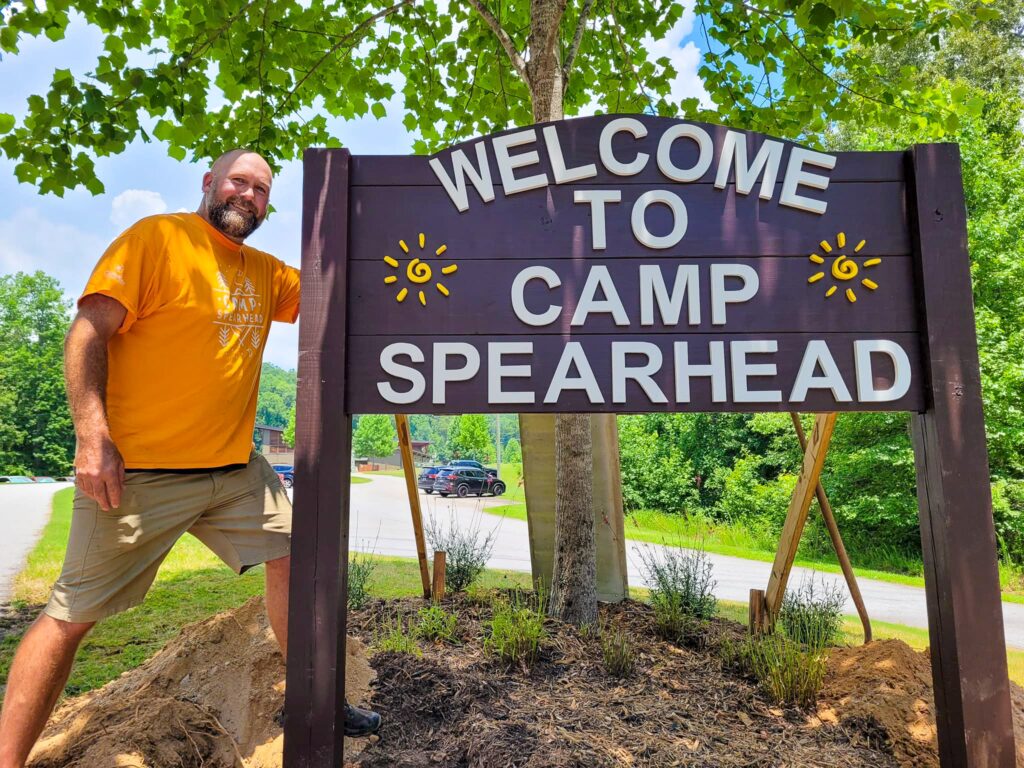 A man standing beside a sign that reads "Welcome to Camp Spearhead".