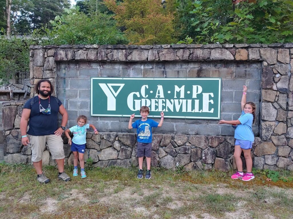 A man and three children pose in front of the YMCA Camp Greenville sign.