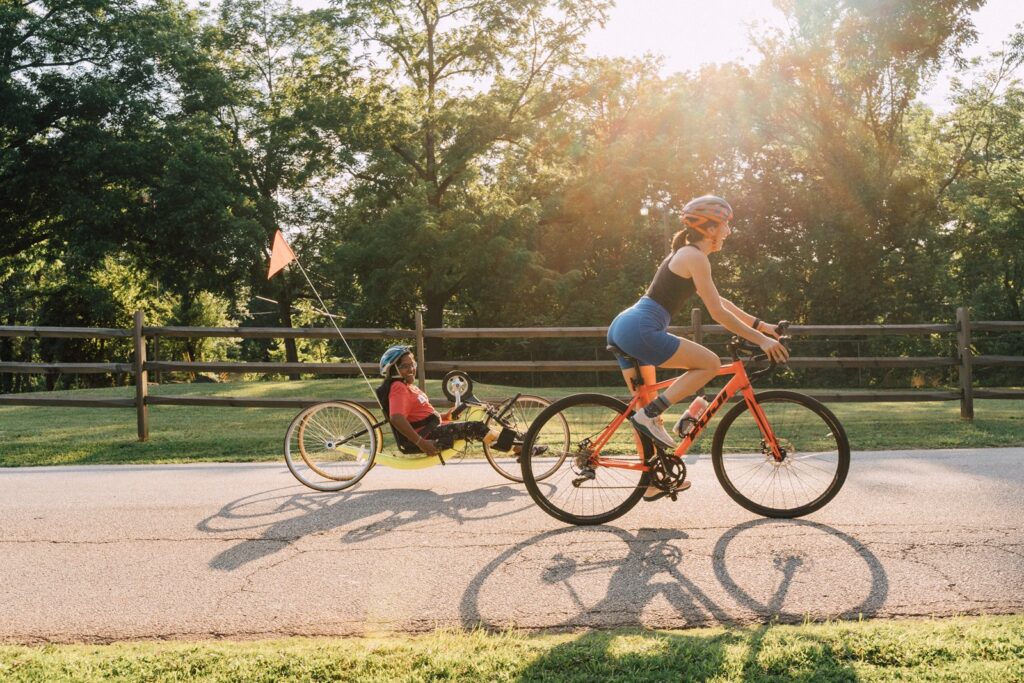 Two women ride bikes. One bike is standard while the other bike is a handcycle.