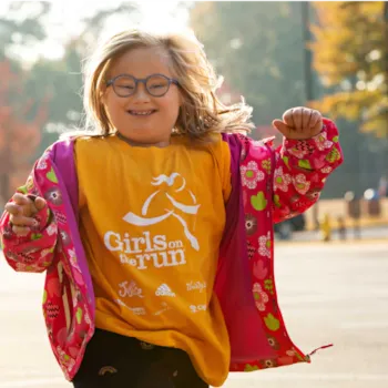 A young girl wearing glasses and a yellow Girls on the Run tshirt smiles as she runs outside.