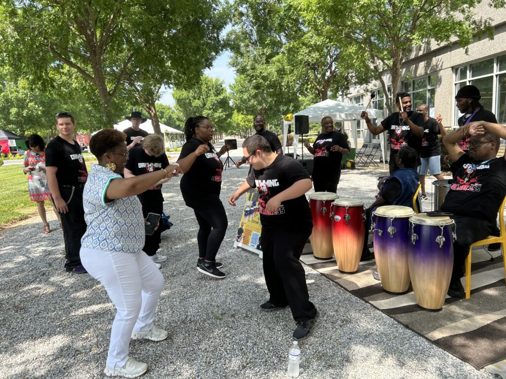 Drummers playing standing drums while people dance and enjoy the music at an outdoor event.