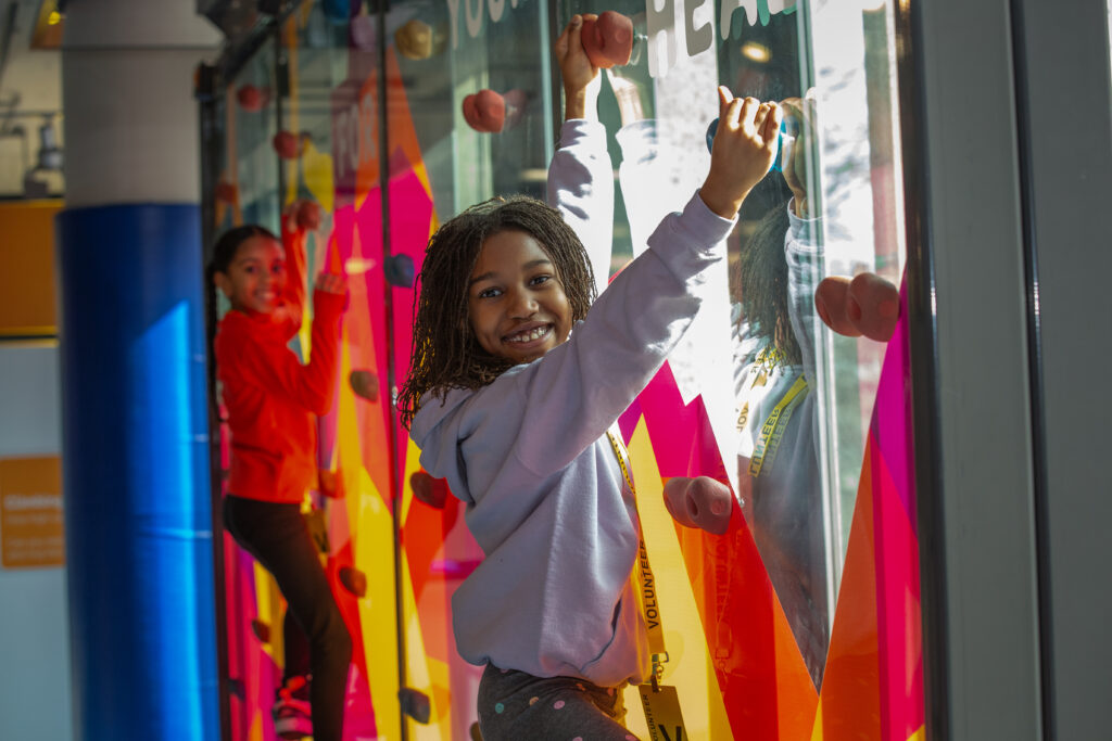 2 young smiling girls are facing the camera and are on a climbing wall.