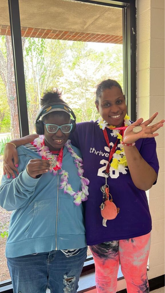 Two women stand side by side with arms around each other, showing the camera their Special Olympic medals that they where on ribbons around their necks.